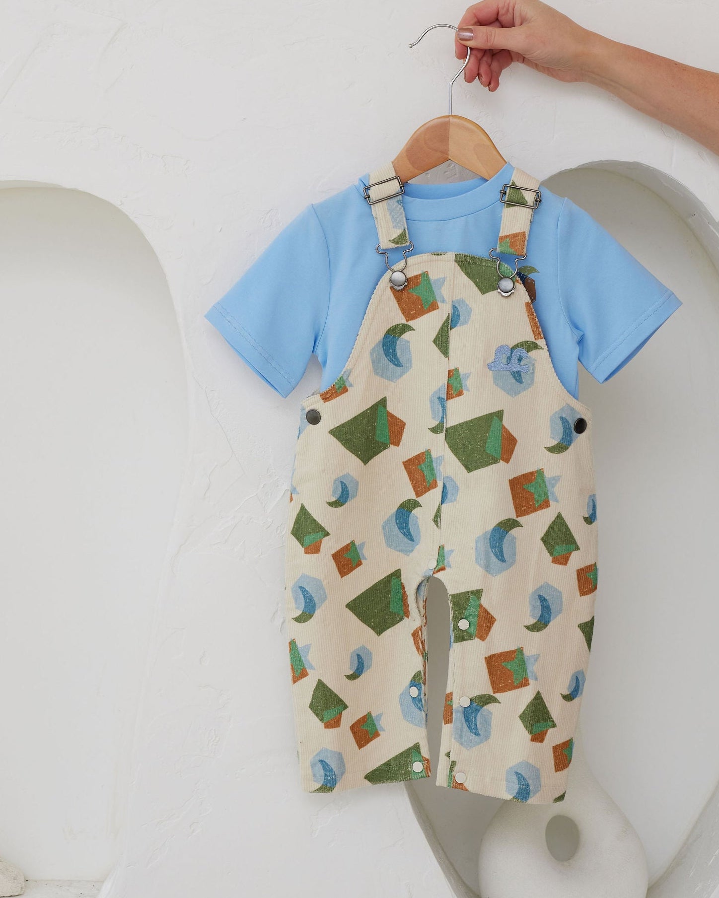 Baby outfit with blue shirt and patterned overalls hanging on a hanger against a white background.