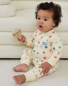 Baby wearing a cream-colored onesie with colorful patterns, sitting on a white floor against a white couch.